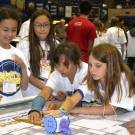 Young students wear white T Shirts with event logo and work on robotics projects indoors