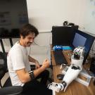 Person sits at a desk wearing a mechanical arm band and looking at a robotic arm