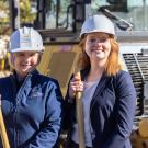 Hawkins and Harvey stand outdoors near construction equipment, wearing hard hats